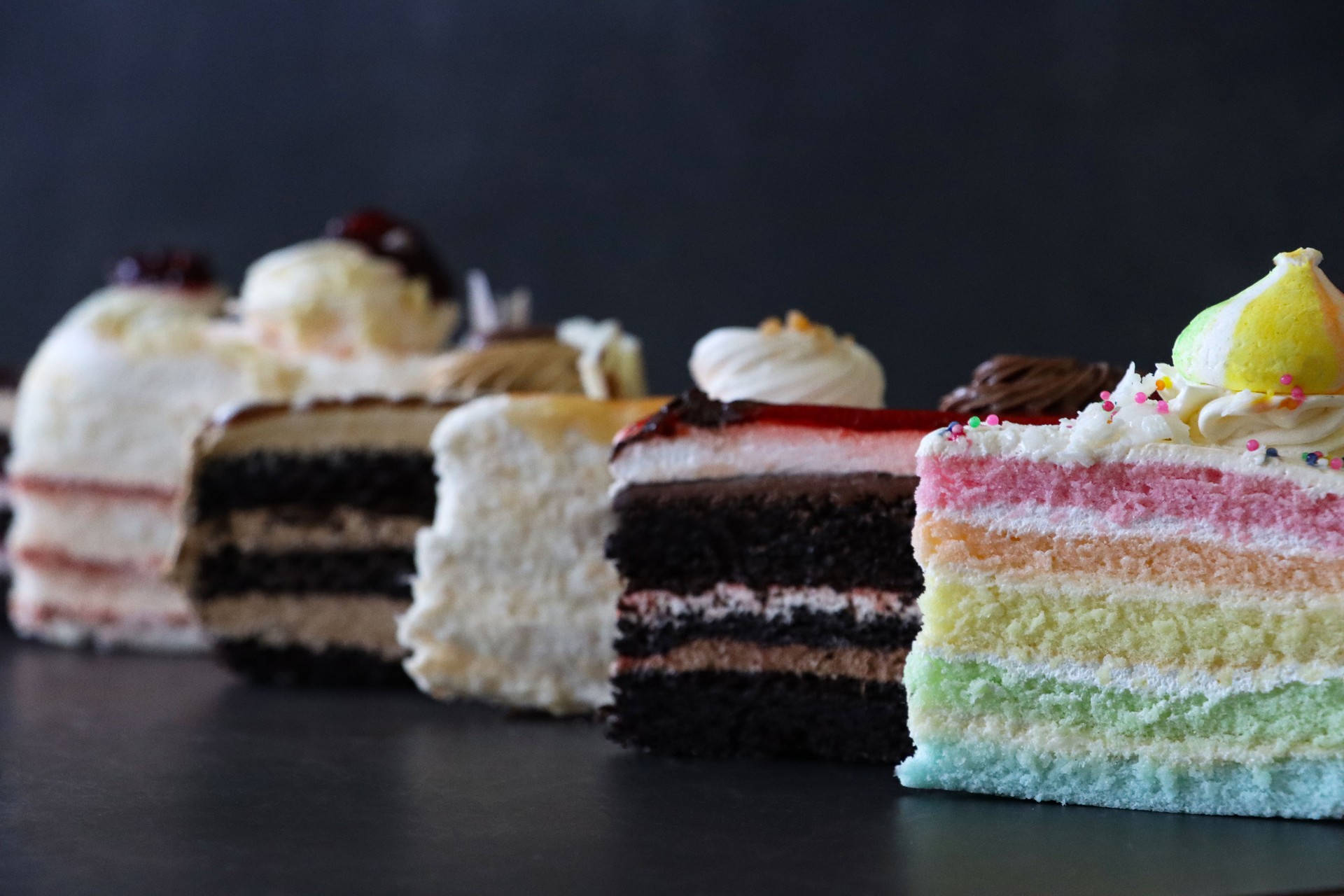 Close-up image of row of six slices of gateaux varieties, Black Forest gateau, angel food cake, coffee and marbled gateau, salted caramel cake, chocolate and cherry cake, rainbow cake, mottled black background, focus on foreground